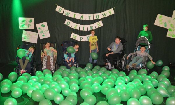 Na zdjęciu widzimy siedem osób. Pięć osób siedzi na wózkach inwalidzkich. Dwie stoją. Wśród nich jest dużo zielonych balonów. Za nimi na ścianie widoczny jest napis: Światowy Dzień MPD.