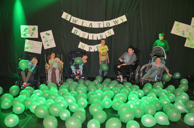 Na zdjęciu widzimy siedem osób. Pięć osób siedzi na wózkach inwalidzkich. Dwie stoją. Wśród nich jest dużo zielonych balonów. Za nimi na ścianie widoczny jest napis: Światowy Dzień MPD.