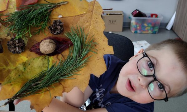 Na zdjęciu widzimy chłopca w okularach, który pokazuje wykonaną przez siebie pracę plastyczną. Jest to stroik jesienny wykonany z liści, gałęzi, szyszek i orzecha.