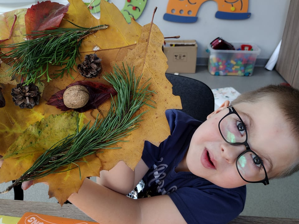 Na zdjęciu widzimy chłopca w okularach, który pokazuje wykonaną przez siebie pracę plastyczną. Jest to stroik jesienny wykonany z liści, gałęzi, szyszek i orzecha.