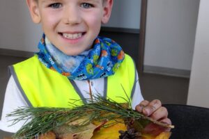 Na zdjęciu widzimy chłopca, który się uśmiecha. W dłoniach trzyma jesienny stroik wykonany z liści, szyszki i dzikiej róży.