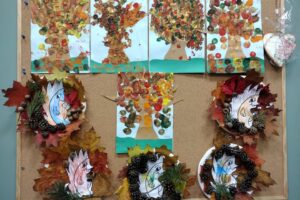 Na zdjęciu widzimy prace plastyczne dzieci, które są przyczepione do tablicy korkowej. Są na niej stroiki jesienne oraz pomalowane farbami obrazy przedstawiające drzewa.