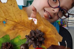 Na zdjęciu widzimy chłopca w okularach, który się uśmiecha. W dłoniach trzyma stroik jesienny wykonany z liści, szyszek i dzikiej róży.