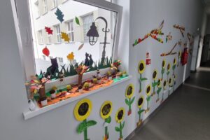 Na zdjęciu widzimy okno i ścianę przyozdobioną pracami plastycznymi dzieci. Są to kwiaty, gałęzie, liście, drzewa w jesiennej aranżacji.
