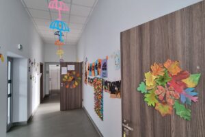 Na zdjęciu widzimy korytarz, który przyozdobiony jest pracami plastycznymi dzieci. Od góry wiszą jesienne papierowe parasole. Na drzwiach przyklejone są kolorowe papierowe liście. Na ścianach wywieszone są prace plastyczne dzieci.