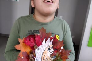 Na zdjęciu widzimy chłopca w okularach, który się uśmiecha. W dłoniach trzyma stroik jesienny. Stoik wykonany jest z liści, szyszek, papieru i żołędzi.