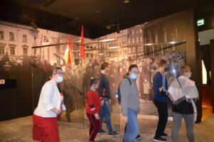 Na zdjęciu widocznych jest 6 osób. Przechodzą przez pomieszczenie w Muzeum Śląskim w Katowicach. Na ścianach widoczne są fototapety ze starych zdjęć.