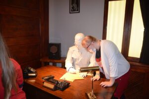 Na zdjęciu widoczna jest 1 osoba. Znajduje się w pomieszczeniu Muzeum Śląskiego w Kawtowicach. Ogląda ekspozycję muzealną, gdzie przy biurku umieszczona jest rzeźba człowieka podpisującego dokumenty.