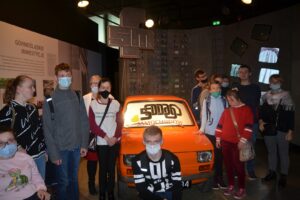 Na zdjęciu widocznych jest 13 osób. Stoją w pomieszczeniu Muzeum Śląskiego w Katowicach. Obok nich znajduje się pomarańczowy samochód.