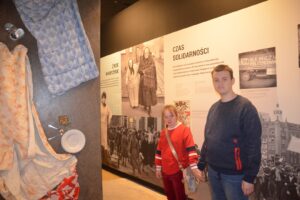 Na zdjęciu widoczne są 2 osoby. Znajdują się w Muzeum Śląskim w Katowicach. Stoją w tle muzealnej ekspozycji, na której widoczny jest tekst i czarno-białe zdjęcia.