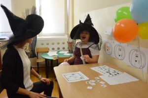 Na zdjęciu widoczne są 2 osoby. Kobieta i dziewczyna mają na głowie kapelusze czarownic. Obok nich znajdują się balony oraz kartki z wydrukowanymi dyniami. Przed dziewczynką na ławce leżą kartki z zadaniami.