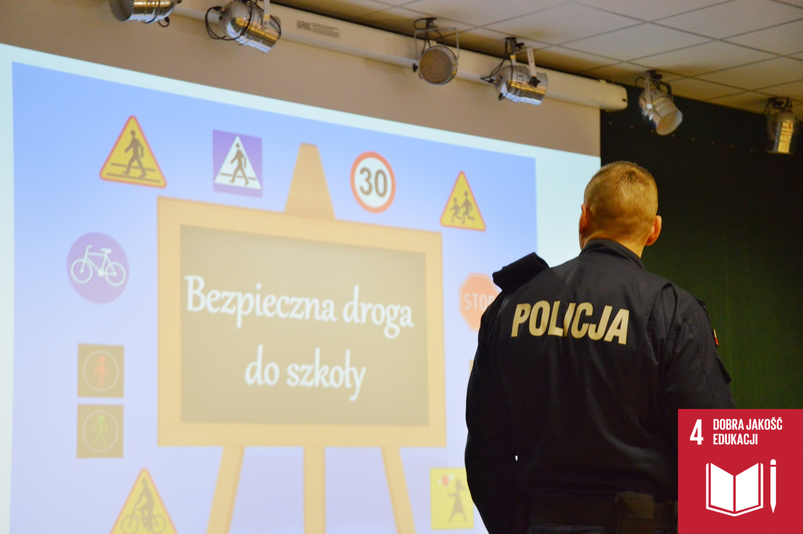 Zdjęcie przedstawia policjanta, który patrzy na wyświetloną za nim planszę. Plansza Przedstawia tablicę z napisem "Bezpieczna droga do szkoły", a wokół tablicy widoczne są znaki drogowe.