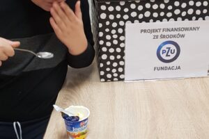 Na zdjęciu widoczny jest chłopiec. Chłopiec siedzi przy stole, trzyma w dłoni łyżeczkę i jedną z dłoni dotyka ust. Przed nim na stole leży jogurt oraz banan i pudełko. Na pudełku widnieje etykieta "Projekt finansowany ze środków PZU Fundacja".
