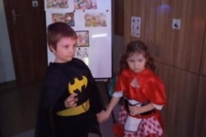 Na zdjęciu widoczne są 2 osoby. Chłopiec przebrany jest za Batmana, dziewczynka za Czerwonego Kapturka. Oboje stoją w sali lekcyjnej i trzymają się za ręce.