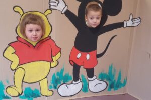 Na zdjęciu widoczne są 2 osoby. Chłopiec i dziewczynka pozują na ściance z postaciami Kubusia Puchatka i Myszki Miki.