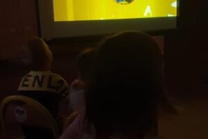 Na zdjęciu widoczny jest ekran, na którym wyświetlana jest bajka. Ekran znajduje się na sali, na której zgaszono światła. Dzieci siedzą na podłodze przed ekranem i oglądają bajkę.