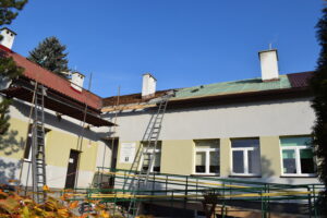 Na zdjęciu widoczny jest fragment budynku. Przed budynkiem rozłożone jest rusztowanie oraz drabiny. Dach budynku jest w trakcie remontu. Przed budynkiem znajduje się żywopłot, drzewa oraz zielony płot.