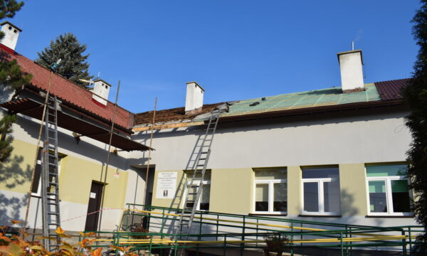 Na zdjęciu widoczny jest fragment budynku. Przed budynkiem rozłożone jest rusztowanie oraz drabiny. Dach budynku jest w trakcie remontu. Przed budynkiem znajduje się żywopłot, drzewa oraz zielony płot.