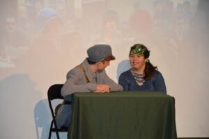Na zdjęciu widoczne są 2 osoby. Chłopiec i dziewczynka. Chłopiec ubrany jest w mundur Polskiego Legionisty, Dziewczynka ma przypięty do włosów zielony kwiat. Oboje siedzą przy stole, który nakryty jest zielonym obrusem.