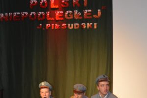 Na zdjęciu widoczne są 3 osoby. Są to chłopcy i znajdują się na scenie. Każdy z nich siedzi po turecku i ubrany jest w mundur Polskiego Legionisty. Chłopiec po lewej stronie trzyma w dłoni mikrofon.
