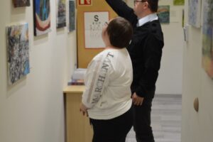 Na zdjęciu znajdują się dwie osoby. Kobieta i mężczyzna stoją bokiem do fotografującego. Oboje patrzą na obrazy zawieszone na ścianie. Mężczyzna wskazuje dłonią na jeden z obrazów.