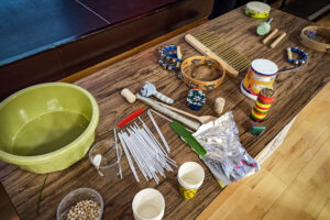 Na zdjęciu widoczny jest stół, na którym położonych jest dużo przedmiotów. Miska z wodą, pojemnik z ziarnami kukurydzy, papierowe kubki, słomki, folia aluminiowa, grzebienie, korek, tamburyna i grzechotki.