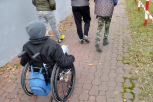Na zdjęciu widocznych jest 5 osób. Na pierwszym planie widzimy chłopca, który jedzie na wózku inwalidzkim. Przed nim chodnikiem spaceruje 4 osoby.