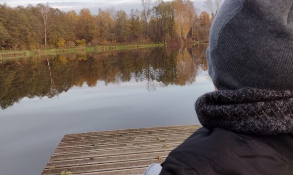 Na zdjęciu widoczna jest 1 osoba. Jest odwrócona tyłem do fotografującego. Patrzy na jezioro znajdujące się przed nim.