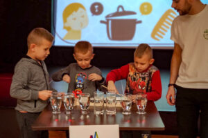 Na zdjęciu widoczne są 4 osoby. 3 chłopców i 1 mężczyzna. Chłopcy stoją obok stolika, na którym ustawione są szklanki z wodą. Obok chłopców stoi mężczyzna i spogląda na nich.