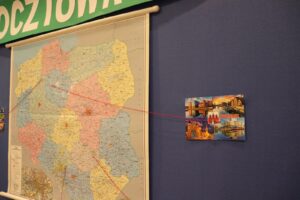 Na zdjęciu widoczna jest tablica z mapą i pocztówka. Pocztówka jest połączona nicią z miejscowością na mapie.