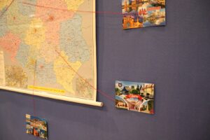 Na zdjęciu widoczna jest tablica z mapą i 3 pocztówkami. Pocztówki połączone są czerwoną nicią z różnymi miejscowościami na mapie.
