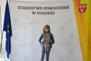 Na zdjęci widoczna jest dziewczynka, która trzyma w ręku dyplom. Stoi na tle ścianki, na której napisane jest Starostwo Powiatowe w Olkuszu.