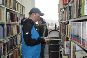 Na zdjęciu 2 uczestników WTZ stoi między regałami wypełnionymi książkami. Pierwszy z nich trzyma książkę w ręku.